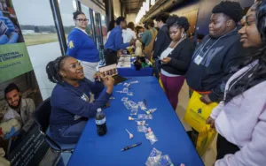 A group of students interacts at a university booth, with a representative speaking and showcasing materials. Colorful items are displayed on a blue table, and visitors hold yellow bags.