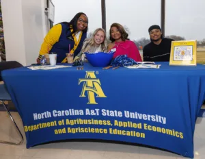 Four students pose at a table featuring North Carolina A&T State University's Agribusiness program. They smile and engage with promotional materials, including a QR code.