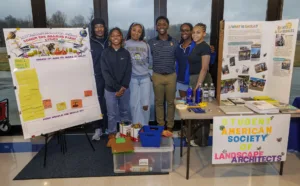 A group of six students poses by a display for the Student American Society of Landscape Architects. The display features a design activity and informational materials, with various tools and projects on the table.