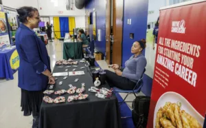 Two women are engaged in conversation at a career fair booth. One woman stands beside a table displaying various promotional items, while the other sits, smiling and holding a paper. A banner promoting job opportunities is in the background.