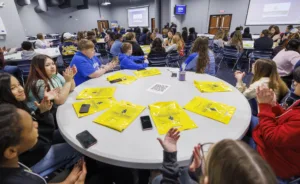 A diverse group of students sits at a round table, some clapping. Yellow bags are placed on the table, and a presentation is visible in the background. The atmosphere is engaged and collaborative.
