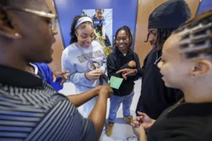 A group of young people smiles and interacts enthusiastically while examining a green card in a school hallway. A poster with a student's image is visible on the wall behind them.