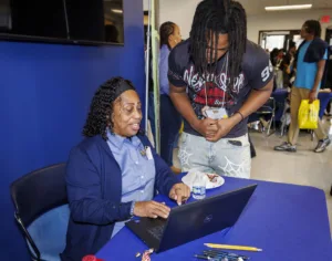 A woman sits at a table smiling while working on a laptop, helping a young man who is leaning over to look at the screen. Other people are visible in the background.