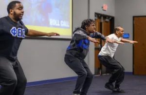 Three young men perform synchronized squat movements in a classroom setting, demonstrating energy and focus. A colorful "ROLL CALL" slide is visible in the background.