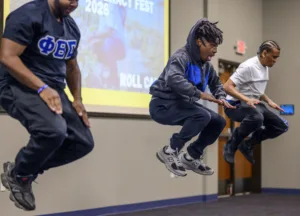 Three individuals in athletic wear are mid-jump during a performance or activity. A presentation screen with "Impact Fest 2026" is visible in the background.