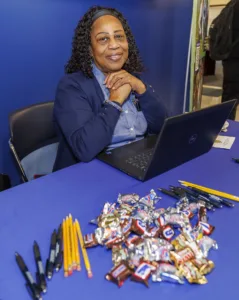A smiling woman with curly hair sits at a table with a laptop, surrounded by candy and pens. Yellow pencils are arranged neatly beside various chocolate candies. The background features a blue wall.
