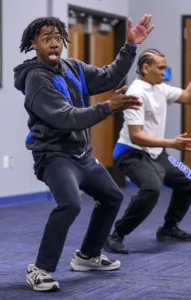 Two dancers are in a studio, performing dynamic moves. One dancer crouches low with an expressive face, while the other stands behind, also engaged in the dance. The setting has blue accents and wooden doors in the background.