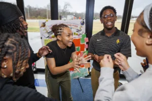 A group of five young adults interact enthusiastically in a bright room, engaged in discussion. A colorful poster is displayed in the background, suggesting a collaborative activity or event.