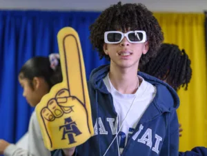 A young person with curly hair and white sunglasses holds a large foam finger while smiling. A person with their back to the camera is visible in the background. The setting features blue and yellow curtains.