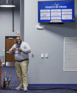 A speaker in a gray sweatshirt presents in a classroom. Behind him is a screen displaying "Points of Pride" with various statistics about a university. A bag is placed on the floor beside the speaker.
