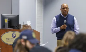 A speaker stands at a podium, addressing an audience with a microphone in hand. The backdrop features a computer and branding for North Carolina A&T State University.
