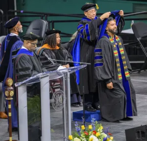 A graduate receiving a hooding ceremony on stage, surrounded by faculty in academic regalia. A podium with a logo is visible, and flowers decorate the area.