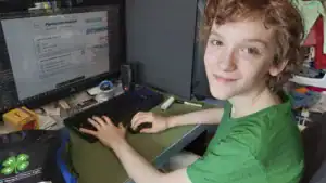 A young person with curly hair sits at a desk, smiling while using a computer. The screen displays a "Planning with Assistant" interface, and various items are scattered around the workspace.
