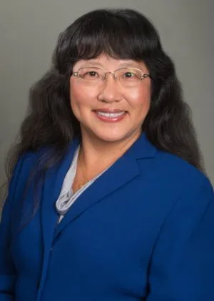 A woman with long black hair and glasses smiles, wearing a blue blazer and light gray blouse, against a neutral background.