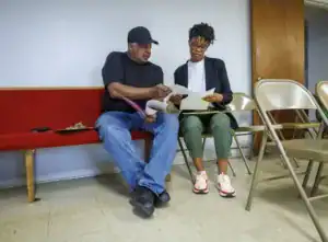 A man and a woman sit on a red bench, discussing documents. The man wears a black cap and t-shirt, while the woman has glasses and is dressed in a blazer, looking at her papers with interest.