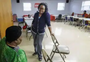 A woman in a navy cardigan stands with a chair in hand while talking to another woman seated nearby in a community center setting. Tables and chairs are arranged in the background.