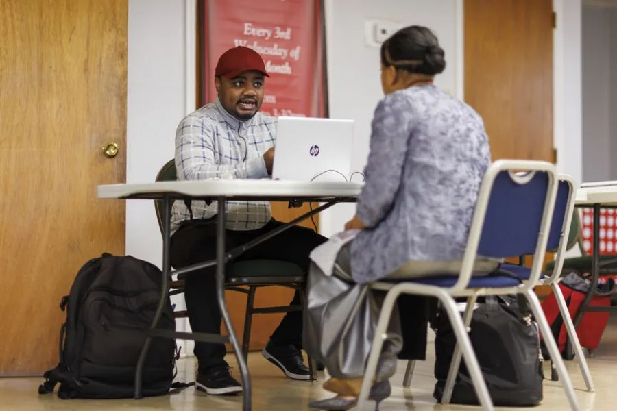 A man in a cap and plaid shirt sits at a table facing a woman in a gray outfit, both engaged in conversation. A laptop is open in front of them, with bags nearby.