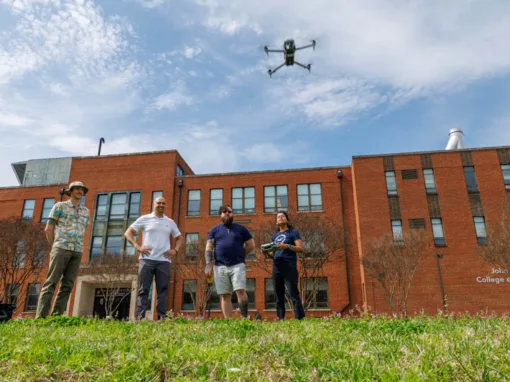 Four people stand on grass in front of a brick building, watching a drone flying above them. One person holds a drone controller, while others observe the flight. The sky is clear with a few clouds.