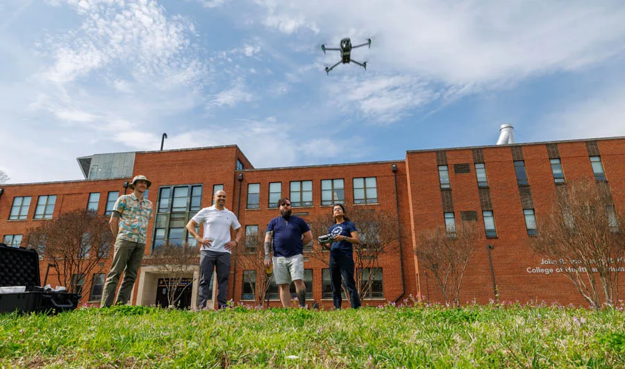 Four people stand on grass in front of a brick building, watching a drone flying above them. One person holds a drone controller, while others observe the flight. The sky is clear with a few clouds.