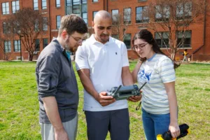 Three people examine a drone controller outdoors in front of a brick building. One man stands between two young adults, who are engaged and pointing at the device while discussing its features.