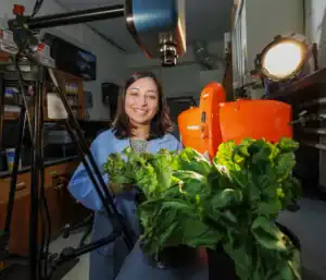 A smiling woman in a lab coat stands next to a camera on a tripod, holding potted lettuce plants. An orange machine and a light source are in the background, indicating a research environment.