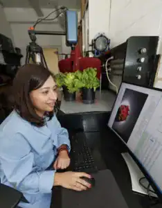 A woman in a lab coat uses a computer to analyze images of lettuce plants, which sit in pots nearby. Various tools and equipment are visible in the background.