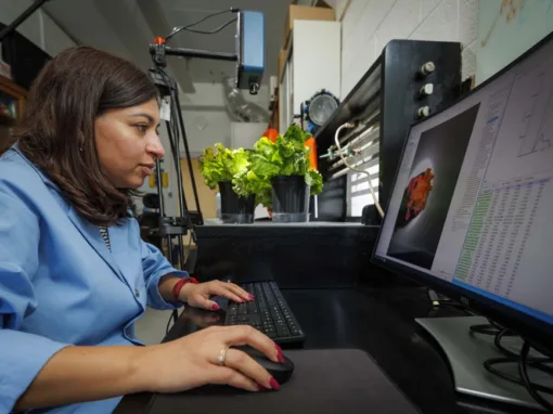 A woman in a lab coat works at a computer, analyzing data. Potted plants are visible in the background, with equipment arranged on the desk around her.