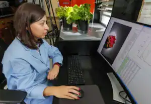A woman in a blue lab coat sits at a computer, focusing on a large monitor displaying colorful data. Two pots of lettuce are visible in the background.
