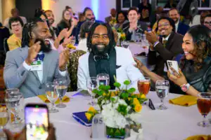 A joyful gathering at a banquet. Three individuals, one in a white tuxedo, share laughter and conversation, while others in the background clap. The table features flowers and drinks, creating a festive atmosphere.