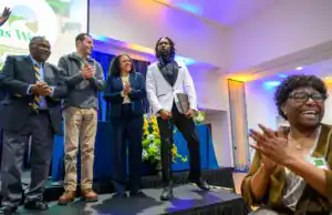 A group of five people applauding during an event. One man in a white suit stands confidently while others smile and clap, showing a celebratory atmosphere. Colorful lighting enhances the setting.
