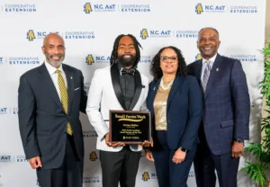 Four people pose together, with one man in a tuxedo holding a plaque that reads 