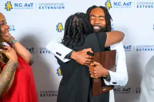 A joyous moment shared between two people hugging, with one holding an award. A third person looks on, smiling. Background features the N.C. A&T State University logo.
