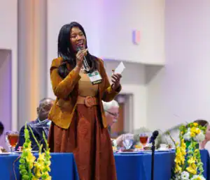 A speaker with long black hair stands at a podium, confidently holding a microphone and notes. She wears a brown blazer and skirt, addressing an audience seated at tables decorated with flowers.