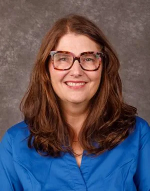 Smiling woman with long brown hair wearing colorful glasses and a blue shirt, set against a gray background.