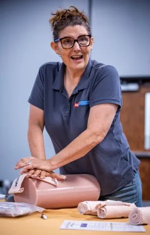A woman with glasses smiles while leaning over a large foam roller on a table. She is wearing a gray polo shirt and is surrounded by rolled towels and a notepad.