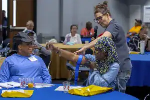 A woman demonstrates a procedure on another woman's arm at a workshop, while an audience observes. Tables are set up with blue tablecloths, and some attendees are engaged in conversation.