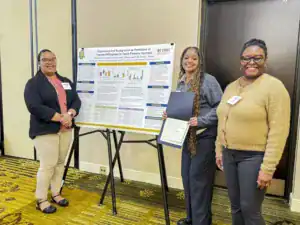 Three women stand in front of a research poster on teaching forestry courses. One holds a certificate, while the others smile at the camera. The background features a neutral-colored wall and a patterned floor.