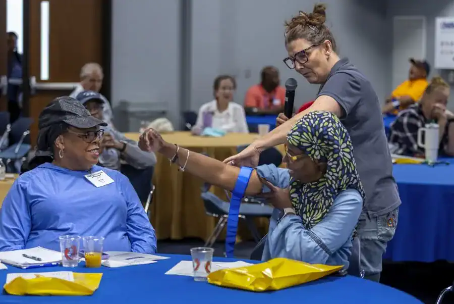A woman demonstrates a procedure on another woman's arm at a workshop, while an audience observes. Tables are set up with blue tablecloths, and some attendees are engaged in conversation.