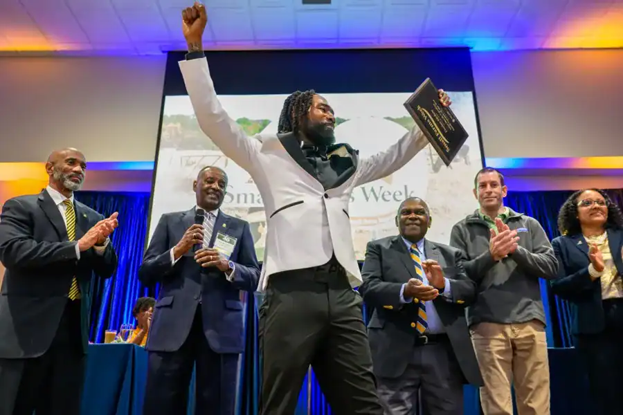 A man in formal attire triumphantly raises a plaque while standing on stage. He is surrounded by five applauding people, including men in suits and women in business attire, with a colorful backdrop and a presentation screen behind them.