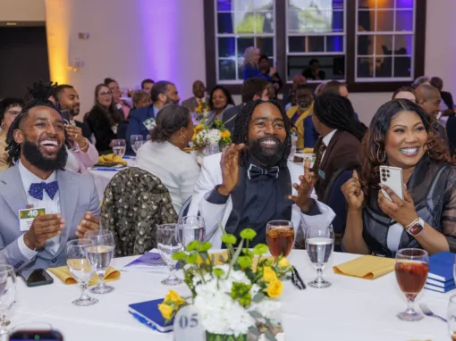 Three smiling guests at a banquet table enthusiastically applauding. The setting features a festive atmosphere with colorful lighting, floral arrangements, and elegantly set tables.