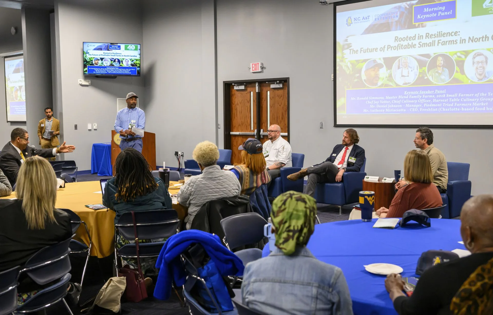 A panel discussion on small farms in North Carolina with several speakers at tables set for an audience. A screen displays the event title and speaker names. Attendees listen attentively in a conference room.