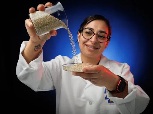 A scientist in a lab coat pours tiny beads from a beaker into a petri dish, smiling. The background features a gradient of blue and red light.