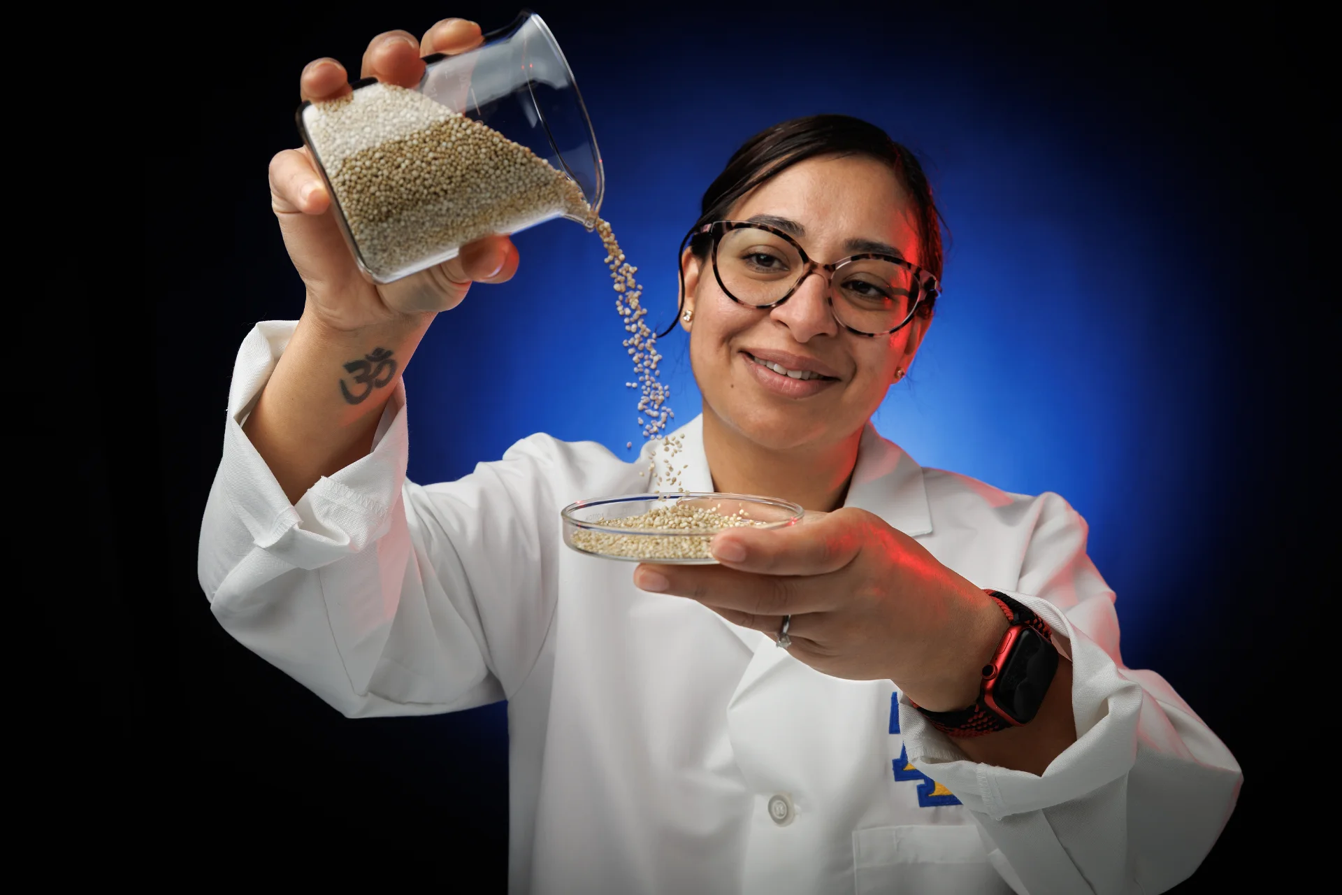 A scientist in a lab coat pours tiny beads from a beaker into a petri dish, smiling. The background features a gradient of blue and red light.
