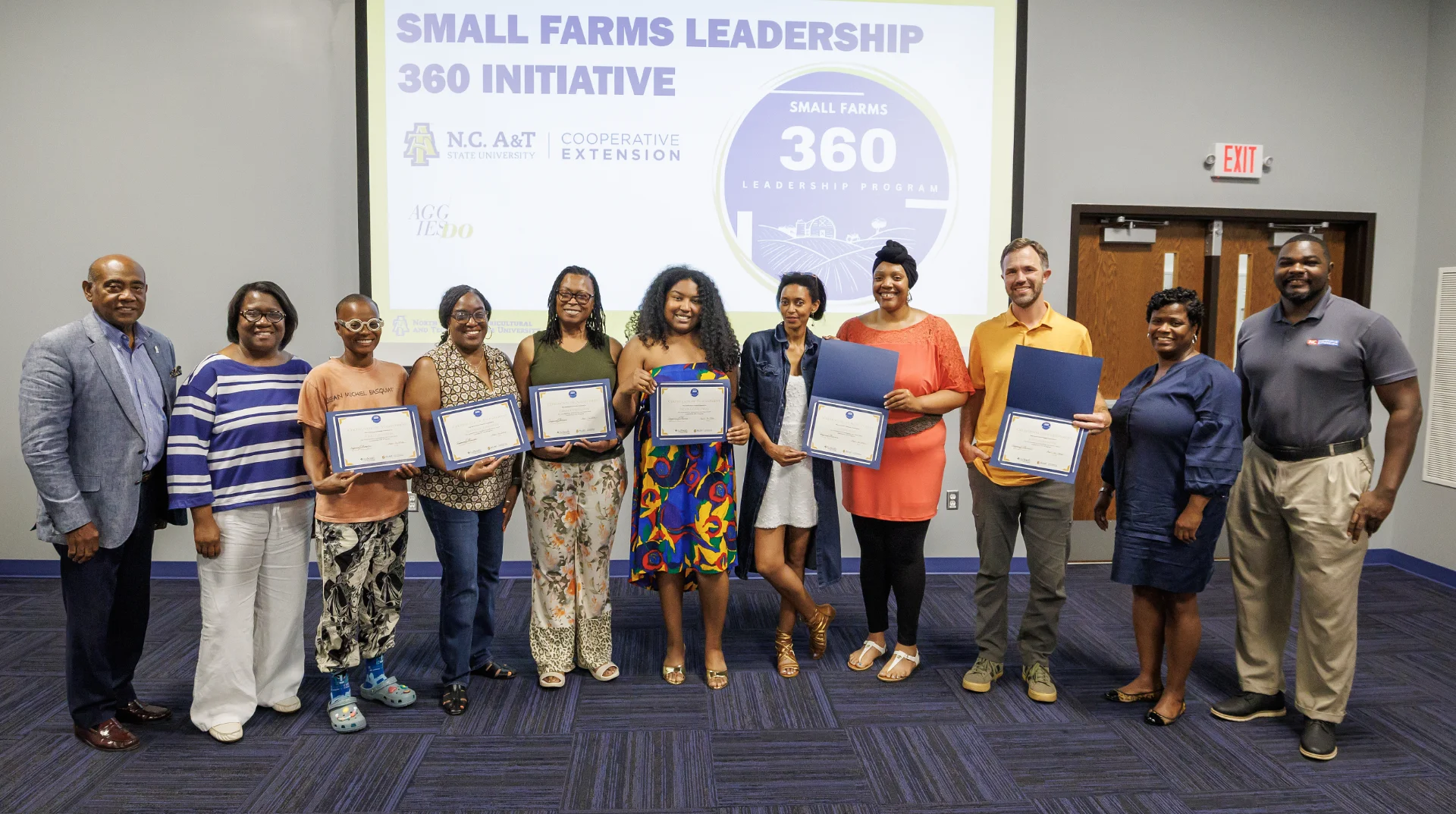 Group of individuals posing with certificates in front of a presentation screen for the Small Farms Leadership 360 Initiative. They are smiling and wearing a variety of clothing styles.