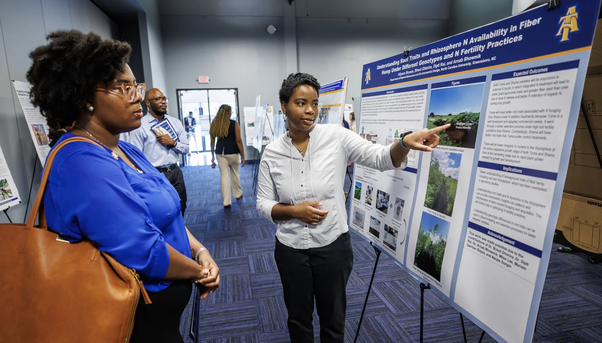 A woman points to a poster about hemp root traits while another woman listens, both engaged in discussion. The setting is an academic presentation with posters on display in the background.