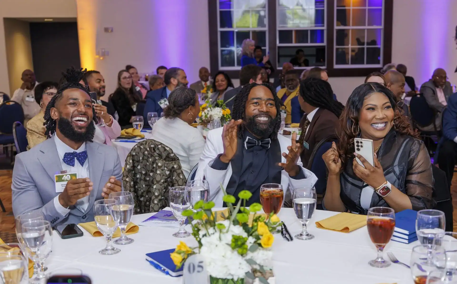 Three smiling guests at a banquet table enthusiastically applauding. The setting features a festive atmosphere with colorful lighting, floral arrangements, and elegantly set tables.