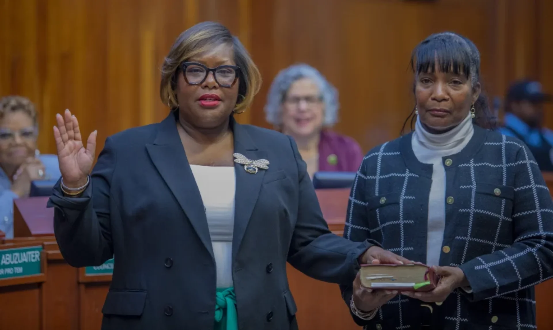 A woman is taking an oath with her right hand raised while holding a book, accompanied by another woman. A group is visible in the background, all in a formal setting.