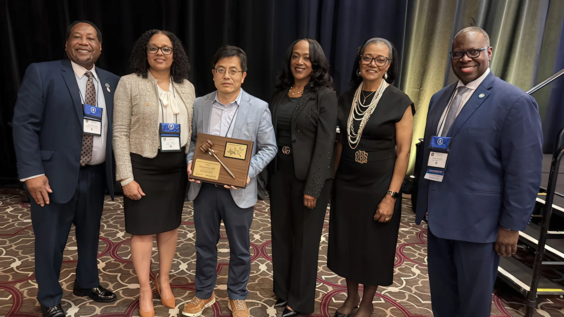 Six people pose together, smiling, at an event. One holds an award plaque. Attendees wear professional attire, with name tags visible. The background features a dark curtain and patterned carpet.