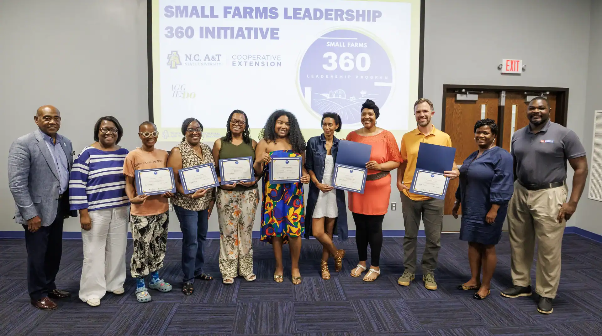 Group of individuals posing with certificates in front of a presentation screen for the Small Farms Leadership 360 Initiative. They are smiling and wearing a variety of clothing styles.