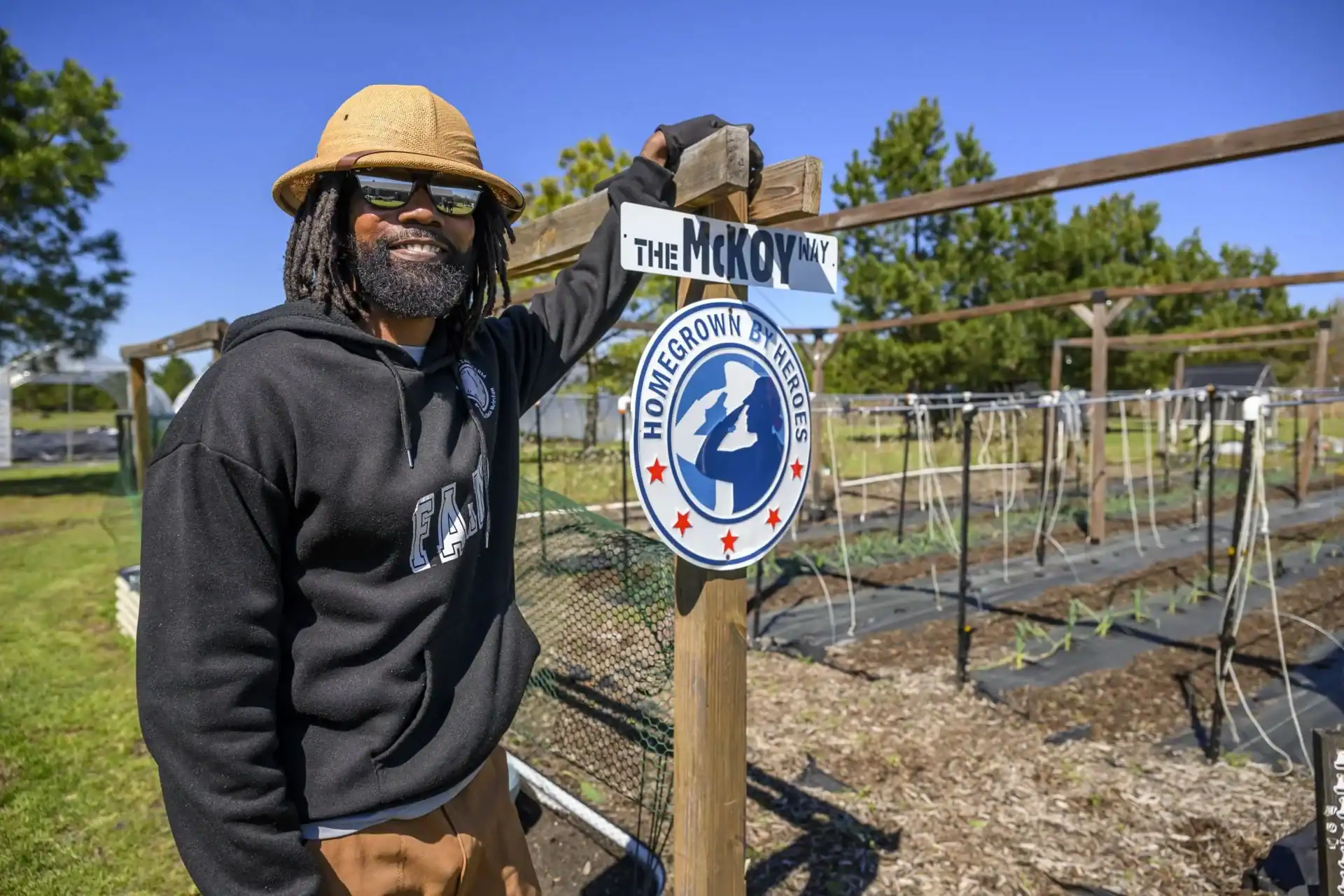 Smiling man with dreadlocks wearing a hat and sunglasses stands by a community garden sign.